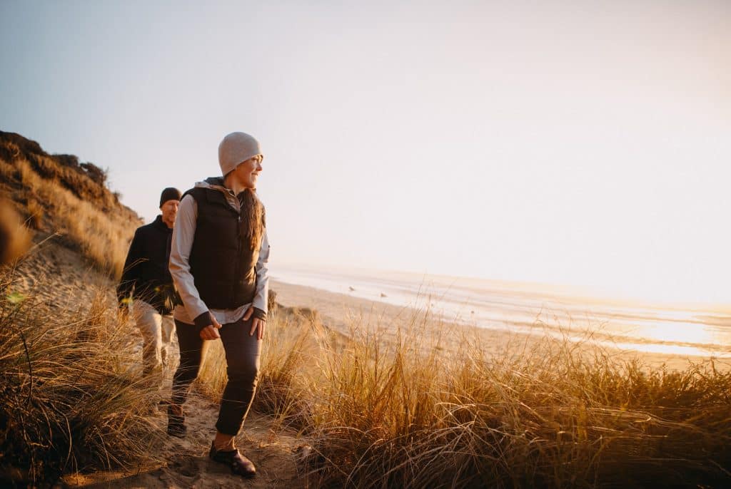 A senior couple explores a beach in Oregon state, enjoying the beauty of sunset on the Pacific Northwest coast.  They hike up a sand dune, the ocean visible in the background.
